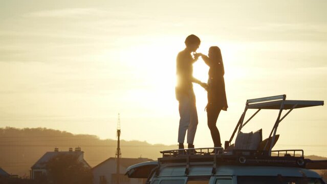 Beautiful view of hipster couple dancing on camper van rooftop, yellow sunset in background. Pair in love enjoying romantic road trip to backcountry. Dark silhouettes, yellow sky