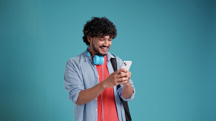 indian or spanish young male in casual with headphones and smartphone use app and smile on blue studio background