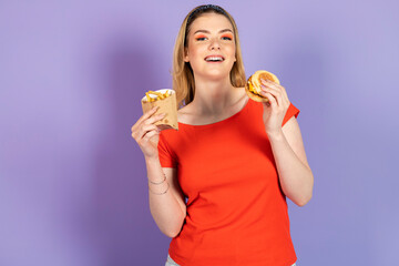 happy smiling pretty girl eats fast food, cheap burger, holding fries and burger. fast food and unhealthy diet. isolated on purple background.