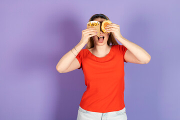 happy pretty girl eats fast food, cheap burger, holding and covering eyes with fries and burger, closed eyes, mouth open. fast food and unhealthy diet. isolated on purple background.