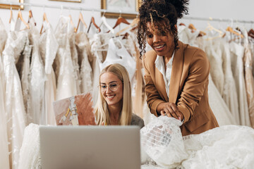 Two women work in a bridal tailoring salon and talking on a video call with a client to choose...