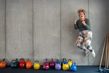 Body positive ecstatic plus-size young woman in fitness clothes jumping for joy in gym feeling confident, satisfied and motivated. Copy space