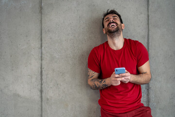 Enthusiastic handsome sports man feeling happy, laughing with eyes closed, standing in gym against the wall, taking a break from workout, holding smart mobile phone.
