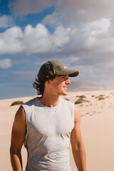 Portrait photo of smiling young man with cap in the desert of the dunes of Corralejo in Fuerteventura, looking at the horizon, in summer, during a vacation trip through the Canary Islands.