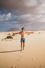 Young man walking happy and shirtless on the desert sand of the dunes of Corralejo in Fuerteventura, in summer, during a vacation trip through the Canary Islands.