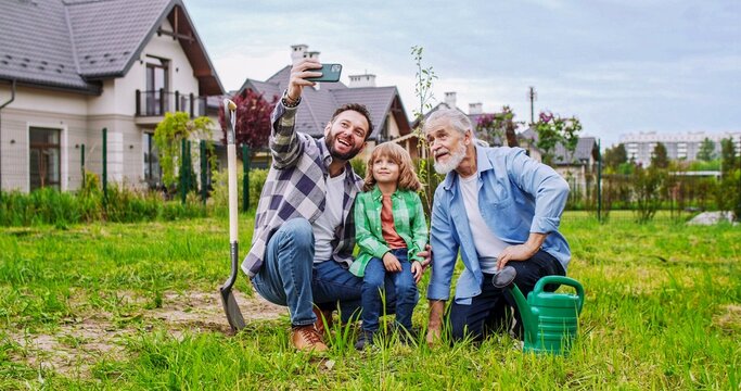 Cheerful man taking selfie photo with smartphone of grandfather, father, son and grandson sitting in garden and smiling to camera. Planting trees concept. Making picture of three generations.