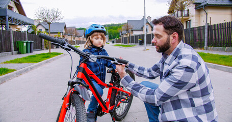 Handsome Caucasian father showing to small cute son in helmet new red bicycle at street. Little boy learning riding a bike. Parenting and care concept. Outdoors.