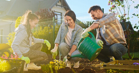 Happy Caucasian family planting flowers or vegetables together in orchard and smiling. Cheerful father watering just planted plants from pot. Sunny summer day. Daughter helping to parents.