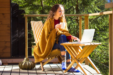 Pretty girl sitting on balcony terrace wrapped in yellow plaid holding cup of tea coffee, looking at laptop. Brunette woman works remotely, reading online. Countryside house. Wood at sunny autumn day.