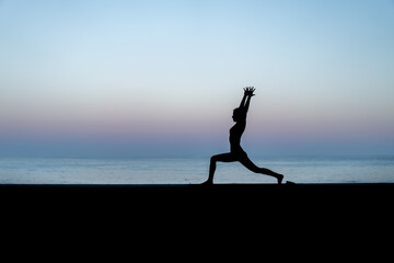Silhouette de femme faisant du yoga devant la mer au coucher de soleil,  posture Virabhadrasana I (Warrior I - Guerrier I)