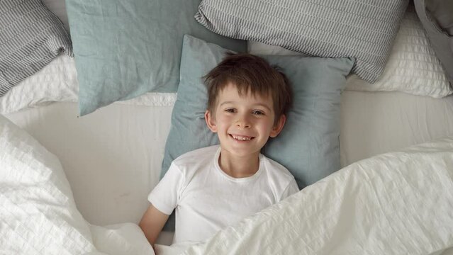 Happy little boy lying in bed and opening his blanket with a smile directed at the camera
