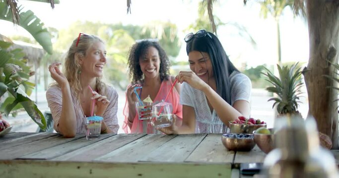 Happy diverse female friends drinking cocktails at beach bar and talking