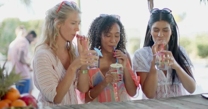Happy diverse female friends drinking cocktails at beach bar and talking
