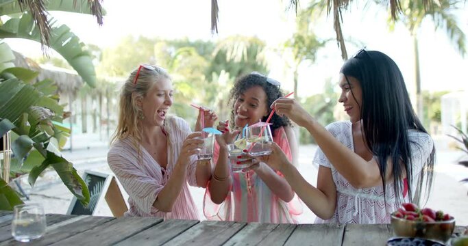 Happy diverse female friends drinking cocktails at beach bar and talking