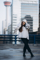 Obraz premium Full length portrait of beautiful brunette smiling girl on the city background. Front view of beautiful girl dressed in white hat and checkered scarf, holding her hat