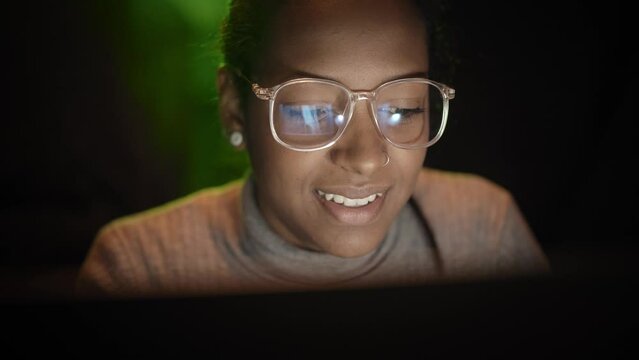 Smiling cheerful mixed race business woman works on computer in dark room. Confident young enterprising girl using laptop computer reading good news on social media. Connected people.