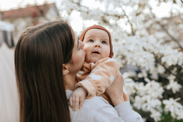 teenage girl with long hair in a grey sweatshirt is holding her newborn sister in a garden with blooming white magnolia.