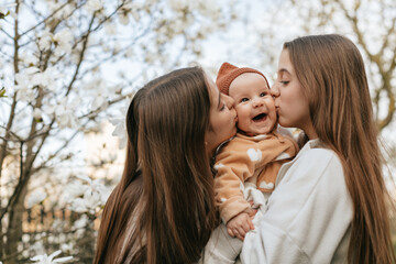 Two teenage girls are holding and kissing a small child against the backdrop of a blooming magnolia. The older twin sisters are kissing the baby's younger sister in a blooming garden.