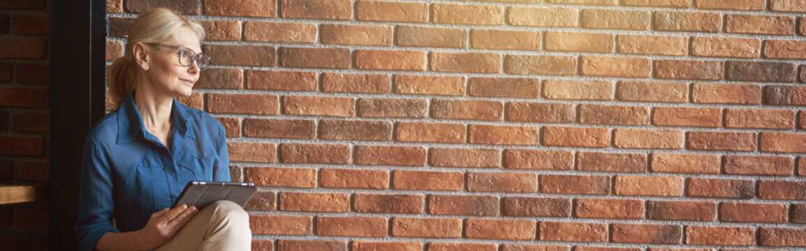 Relaxed Mature Blonde Caucasian Woman Wearing Glasses Looking Away, Using Tablet Pc While Sitting Comfortably On Wooden Stairs In Front Of Brick Wall At Home