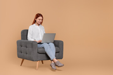 Fototapeta na wymiar Smiling young woman working with laptop in armchair against beige background, space for text