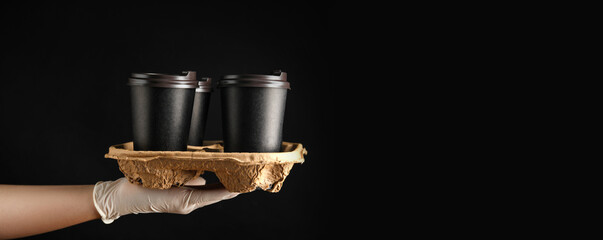 Woman holding cardboard holder with takeaway paper coffee cups on black background, closeup. Banner...