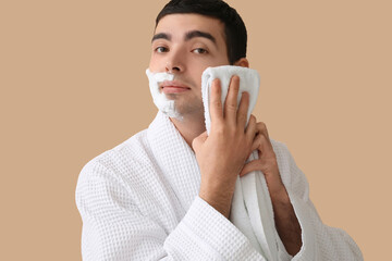 Young man wiping shaving foam from face against beige background