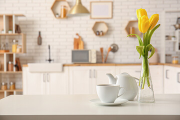Fototapeta na wymiar Cup, teapot and vase with beautiful tulip flowers on table in interior of modern kitchen