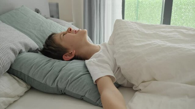 Portrait of smiling boy waking up in bed at morning, yawning and stretching out hands