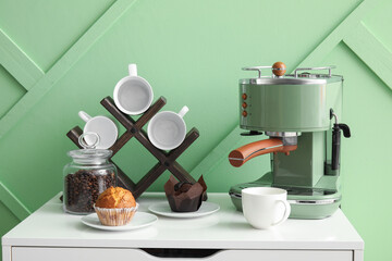 Modern coffee machine, muffins, cups and jar with beans on drawer near green wall