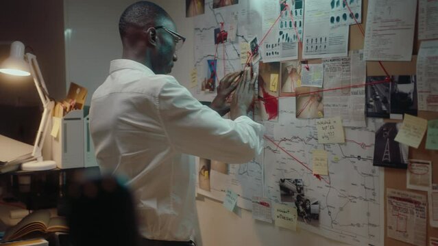 African American male detective pinning crime scene photo to board on the wall, examining notes and clues connected by red string, investigating murder in his office at night