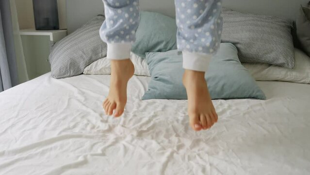 Closeup slow motion of boy's feet jumping up high on the soft bed at bedroom. Happy childhood, fun and joy