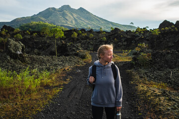 Fototapeta na wymiar A young girl traveler with a backpack walks in the mountains against the backdrop of the Batur volcano on the island of Bali in Indonesia.