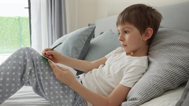 Little boy lying in bed, writing in notebook and ding homework. Concept of education, child development and remote school at home