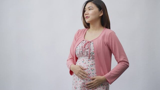 Happy young Asian woman in maternity clothes rubbing her belly while standing in front of a plain white background