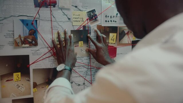 Black detective attaching crime scene photo to pinboard on the wall in his office, looking at clues connected with red thread, working on murder investigation