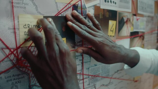 Black detective attaching crime scene photo to pinboard on the wall with pictures, map, notes and fingerprints linked with red thread, investigating murder. Close-up shot