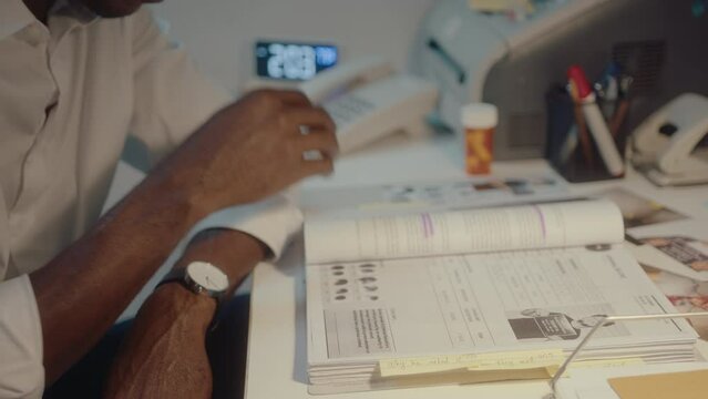 African American criminal investigator sitting at desk, turning pages of crime scene report, reading and analyzing information about murder, working during night in his office