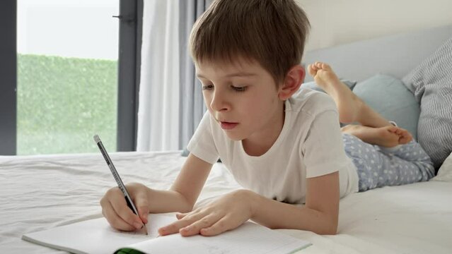 Portrai of little boyworking on his homework in bed, writing in a notebook with concentration. Importance of education and child development in a remote learning environment