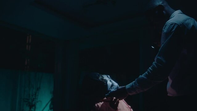 Black detective walking with torch to strangled victim tied to chair with plastic bag on head, checking pulse on neck, working at crime scene at night in dark home with flickering police car lights