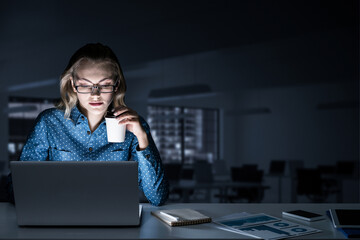 Attractive blonde working on laptop in dark office. Mixed media