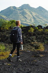 A young girl traveler with a backpack walks in the mountains against the backdrop of the Batur volcano on the island of Bali in Indonesia.