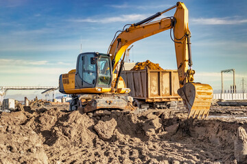 A wheeled excavator loads a dump truck with soil and sand. An excavator with a high-raised bucket...