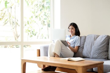 female sitting on sofa and listening to lecture via video call. Attractive young woman in headphones with microphone using laptop, consulting with teacher and taking notes.