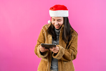 Beautiful Asian girl in a long-sleeved shirt and a Christmas hat. holding a tablet in hand and expressing joy in ordering online products at a discounted price Indoor studio isolated  pink background