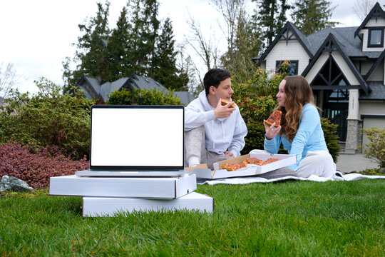 fast fresh pizza delivery youth young brother and sister sitting on picnic eating pizza in background in foreground laptop with blank screen stands on white boxes with delivery summer ad