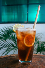 A glass with a refreshing, cold iced tea in front of a mint white wall and tropical plant. Orange brown ice tea with ice cubes and a slice of lemon as decoration on a wooden table.