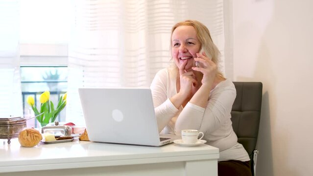 middle-aged adult woman working at computer talking on phone laughing drinking tea coffee light live communication morning breakfast Sun outside window flowers spring pleasant conversation easy life