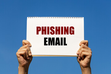 Phishing email text on notebook paper held by 2 hands with isolated blue sky background. This...
