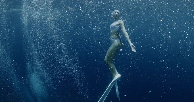 Freediver woman with fins glides underwater in ocean with air bubbles. Freediving water sport in tropical blue sea.