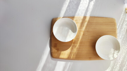 Cutting board made of natural bamboo and white plate saucer on table with shade and light from sun. Abstract natural background, texture, frame, copy space. Concept of cooking with beautiful things